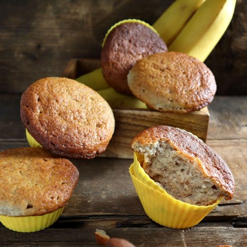 Bolinhos de banana assados em forminhas, com bananas ao fundo sobre mesa de madeira